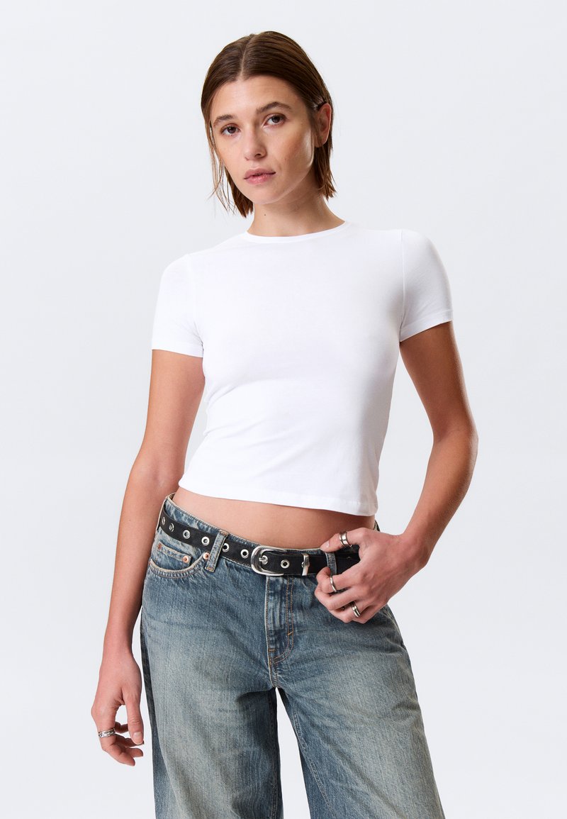 T-shirt blanc ajusté à manches courtes en coton lisse, accompagné d'un jean en denim bleu délavé et ample, avec une ceinture noire à boucle circulaire.