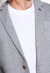 Persona con un blazer gris que lleva un pin de ancla en la solapa sobre una camiseta blanca de cuello redondo, mostrando parte del cuello y el área del pecho.