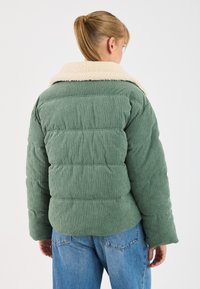 Woman with blonde hair in a ponytail wearing a green corduroy puffer jacket with a cream fleece collar and blue jeans, facing away.