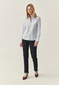 Camisa de botones azul claro de mangas largas, combinada con jeans de mezclilla oscuros. El modelo lleva puestos unos zapatos de tacón marrones. Fondo neutro.