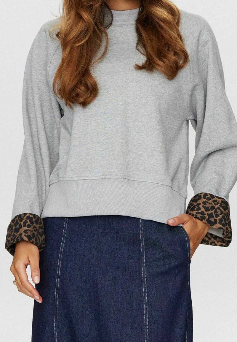 Femme portant un sweat-shirt gris clair avec des poignets à imprimé léopard et une jupe en jean bleu foncé, une main dans la poche, avec des cheveux ondulés lâches visibles.