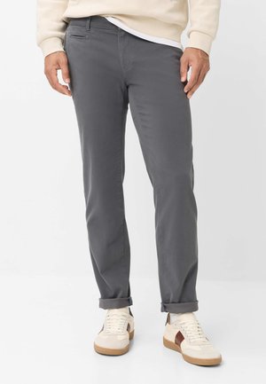Mand iført grå, slanke bukser med opkruede manchetter og hvide sneakers med brune accenter, stående mod en ensfarvet hvid baggrund.