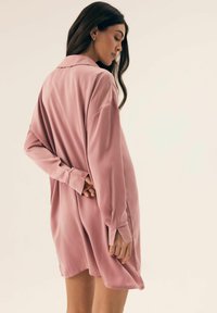 Różowa satynowa sukienka koszulowa z kołnierzykiem, długimi rękawami i luźnym fasonem. Gładka tekstura z bocznymi rozcięciami i mankietami na guziki umożliwiającymi regulację długości.