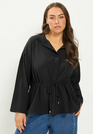 Vrouw met lang bruin haar die een zwarte blouse met lange mouwen en een koord in de taille draagt, met een blauwe spijkerbroek tegen een effen lichte achtergrond.