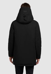 Czarna, ocieplana parka z wysokim kołnierzem i regulowanym kapturem. Charakteryzuje się minimalistycznym designem, prostym krojem i kieszeniami na bokach zapinanymi na zamek.