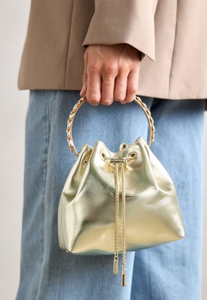 Hand houdt een metalen gouden handtas met een ronde handgreep en trekkoordsluiting, draag een beige jas en blauwe spijkerbroek.