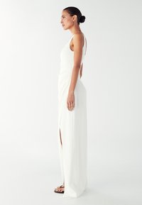 Žena stoji bočno, odjevena u bijelu haljinu bez rukava s bočnim prorezom i crnim sandalama, na jednostavnoj bijeloj pozadini.