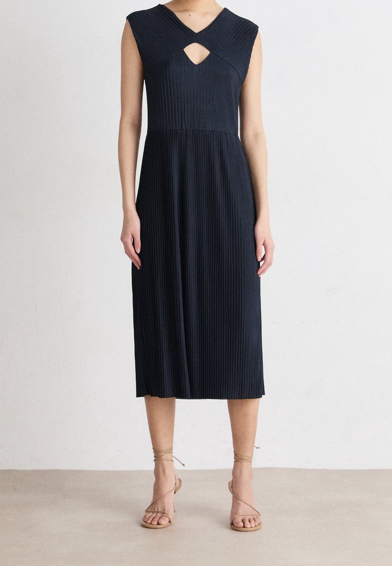 Femme portant une robe midi noire côtelée sans manches avec une petite découpe à l'avant et des sandales beiges à brides.
