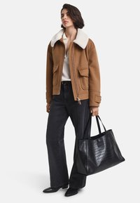 Vrouw in een bruin jasje met een witte kraag, zwarte wijdvallende jeans, zwarte puntige schoenen, en houdt een grote zwarte getextureerde totebag vast.
