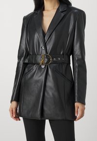 Zwarte leren blazer met een scherpe revers, taillerende taille met een gouden riem met unieke hardware, zijzakken en een gladde textuur.