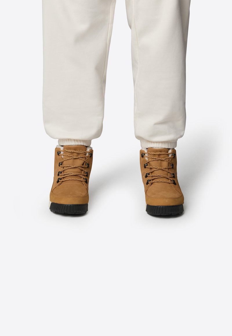Bottes de randonnée marron en cuir texturé, semelles en caoutchouc noir et œillets en métal. Les lacets sont tissés et bouclés, assortis à un pantalon de jogging blanc.