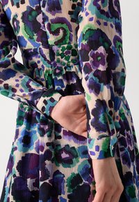 Robe midi florale multicolore avec texture plissée, présentant des nuances de violet, vert et bleu. Manches à revers avec détail en boutonnage.