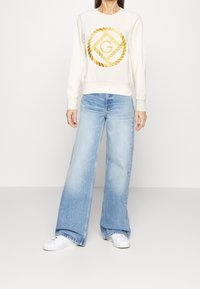 Krämfärgad sweatshirt med en guldbroderad logotyp, kombinerad med högmidjade ljusblå vida jeans och vita sneakers. Enkelt design.