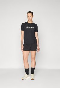 Μαύρο αθλητικό T-shirt με το λογότυπο "Normal", συνδυασμένο με μαύρα σορτς. Μοντέλο που φοράει μπεζ αθλητικά παπούτσια και μαύρες κάλτσες. Απλός, λειτουργικός σχεδιασμός.