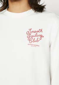 Personne portant une chemise blanche avec un texte rouge indiquant "Smooth Sailing Club" et "Scotch & Soda Amsterdam" sur la poitrine.