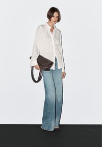 White button-up shirt, light blue flared jeans, and a dark brown woven clutch with a braided handle. Smooth fabric and relaxed fit.