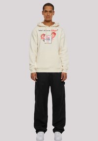 Cream sweatshirt with a hood, featuring a front graphic of children, vintage cereal box, and text. Paired with black pants and white sneakers.