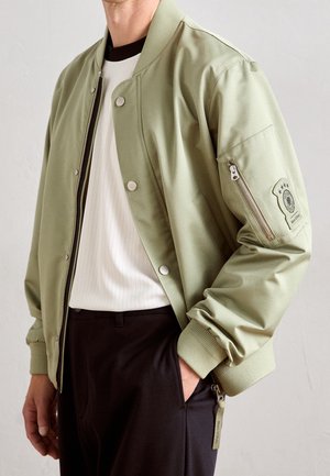 Homme portant une veste bomber vert clair sur une chemise blanche avec un pantalon noir, main dans la poche, sur un fond clair uni.