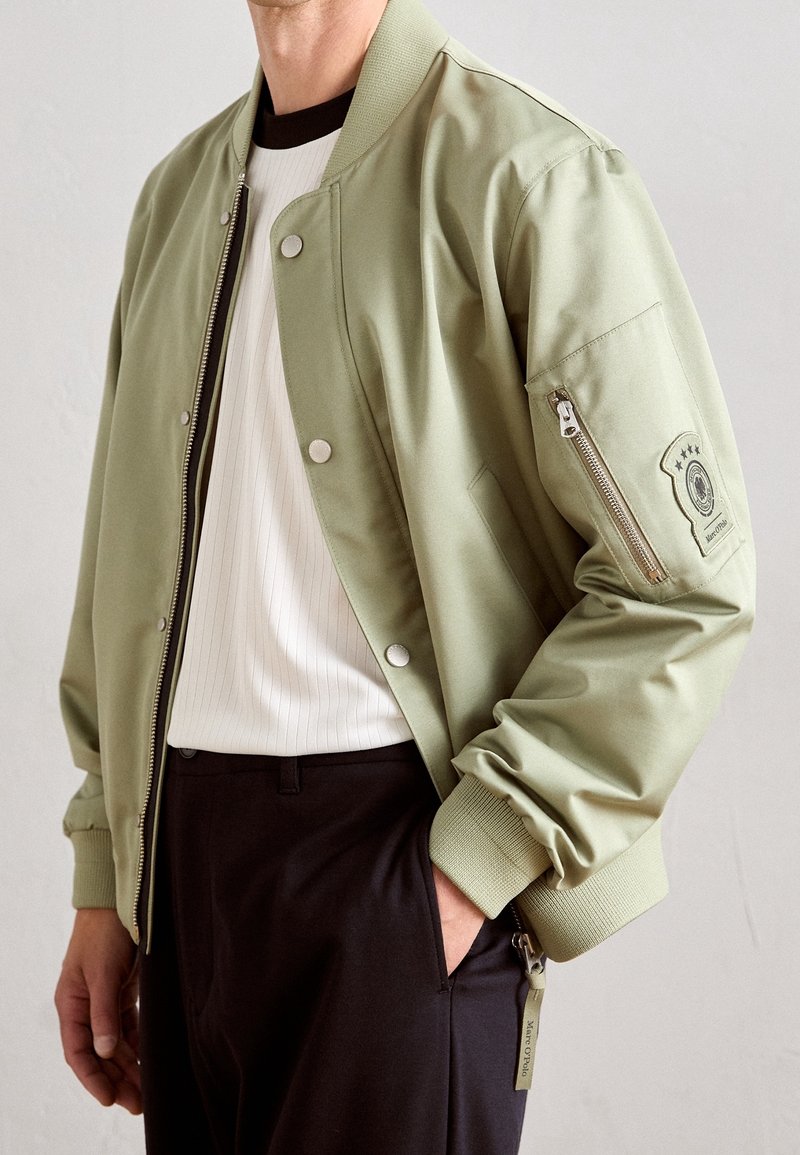 Homme portant une veste bomber vert clair sur une chemise blanche avec un pantalon noir, main dans la poche, sur un fond clair uni.
