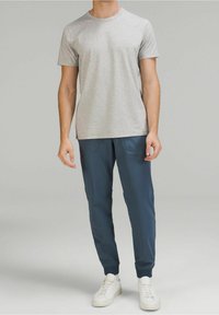 Graues T-Shirt mit kurzen Ärmeln kombiniert mit einer navy Jogginghose mit zwei Vorderaschen. Accessoires umfassen weiße Turnschuhe. Glatte Stofftextur.