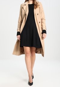 Beige trenchcoat met een dubbele knoopsluiting, zwarte jurk eronder en zwarte hoge hakken. Gladde stof, knielang coat, zonder patronen.