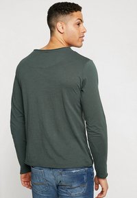Langärmliges Shirt aus dunkelgrünem Stoff, mit Rundhalsausschnitt und glatter Textur. Getragen mit distressed blauen Jeans.