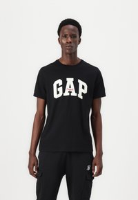 Fekete pamut póló fehér "GAP" felirattal, piros részletekkel. Klasszikus környakú fazon, rövid ujjak és laza szabás.