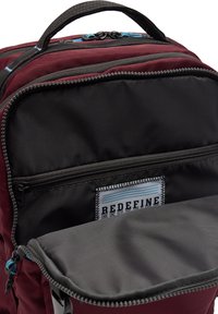 Bordowy plecak z wieloma przegrodami, czarną podszewką, szarym kieszonką i niebieskimi suwakami. Posiada prostokątną metkę wszytą w środku.