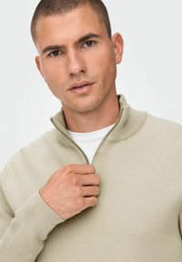 Suéter beige acanalado con cremallera y cuello alto, con una cremallera plateada, llevado sobre una camiseta blanca. La textura es suave y de punto.