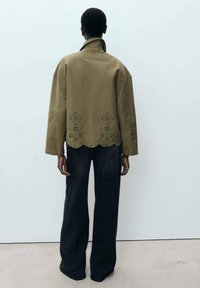 Olijf groene jas met een hoge kraag en een gegolfde zoom, voorzien van kantachtige uitsparingen aan de achterkant. In combinatie met brede zwarte broeken.