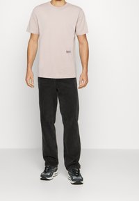 Camiseta de algodón de color rosa claro con mangas cortas, que presenta un logotipo negro impreso en la parte inferior izquierda, combinada con pantalones de mezclilla oscuros y zapatillas.
