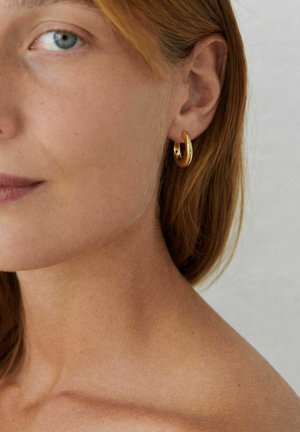 Zbliżenie osoby o jasnej karnacji i rudych włosach, noszącej małą, gładką złotą kolczyk w kształcie koła w lewym uchu.