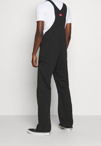 Salopette in denim nero con spalline regolabili, grande tasca frontale e due tasche posteriori. Indossata sopra una t-shirt bianca e abbinata a sneakers nere.