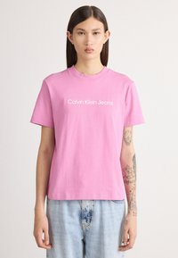 Rozā īsām piedurknēm t-krekls, izgatavots no kokvilnas, ar baltu tekstu "Calvin Klein Jeans" priekšpusē. Brīvs piegriezums, apvienots ar gaiši ziliem džinsiem.