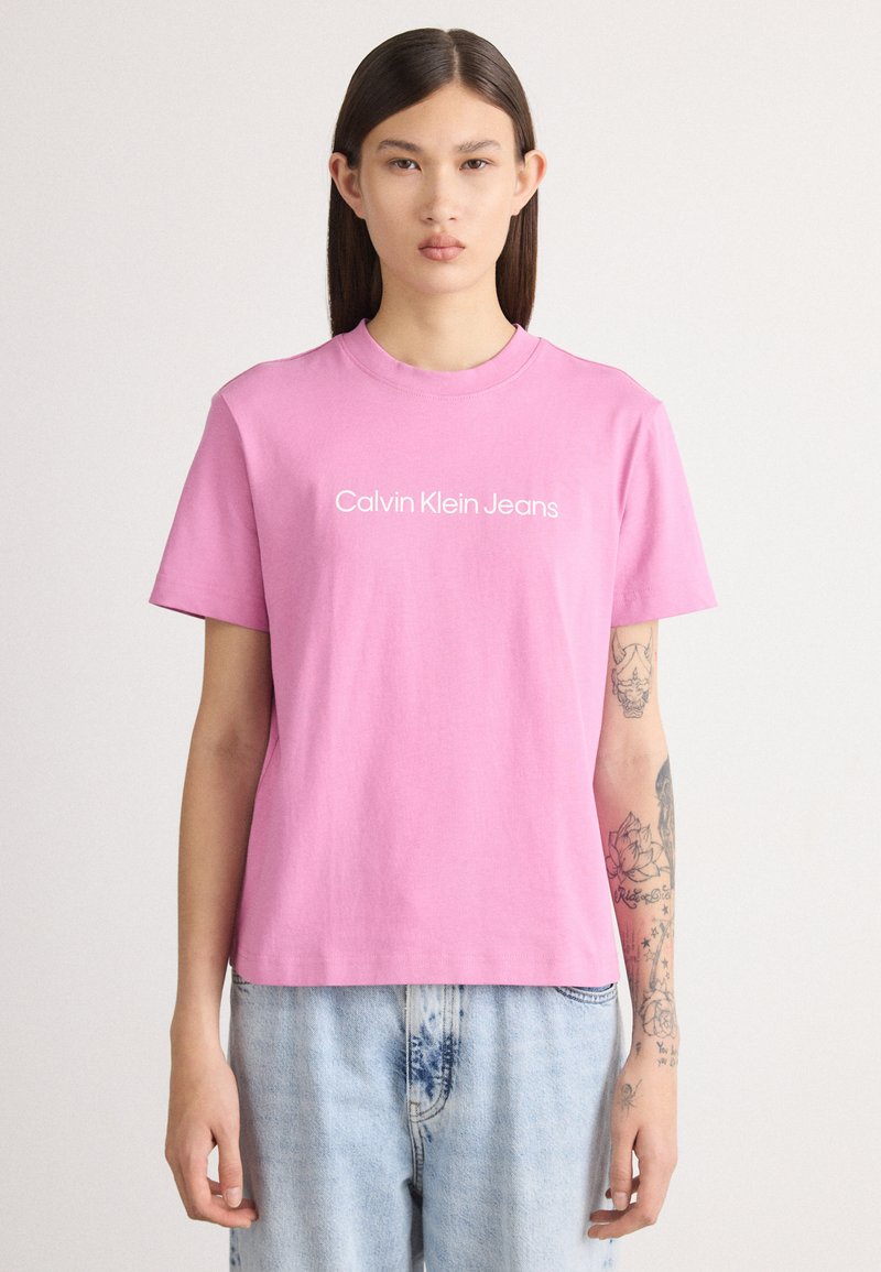 Rozā īsām piedurknēm t-krekls, izgatavots no kokvilnas, ar baltu tekstu "Calvin Klein Jeans" priekšpusē. Brīvs piegriezums, apvienots ar gaiši ziliem džinsiem.