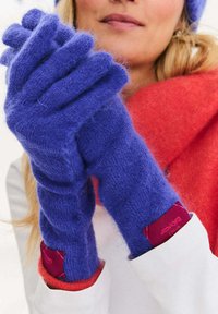 Fialové vlněné rukavice s měkkou texturou a bezprstým designem. Výrazná červená značková visačka na zápěstí. V pozadí je červená šála.