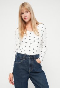 Λευκή μπλούζα με μακρύ μανίκι και μικρό σκούρο λουλούδι σχέδιο, ραφιναρισμένη υφή; συνδυασμένη με ψηλόμεσα σκούρα μπλε τζιν με μπροστινές τσέπες.