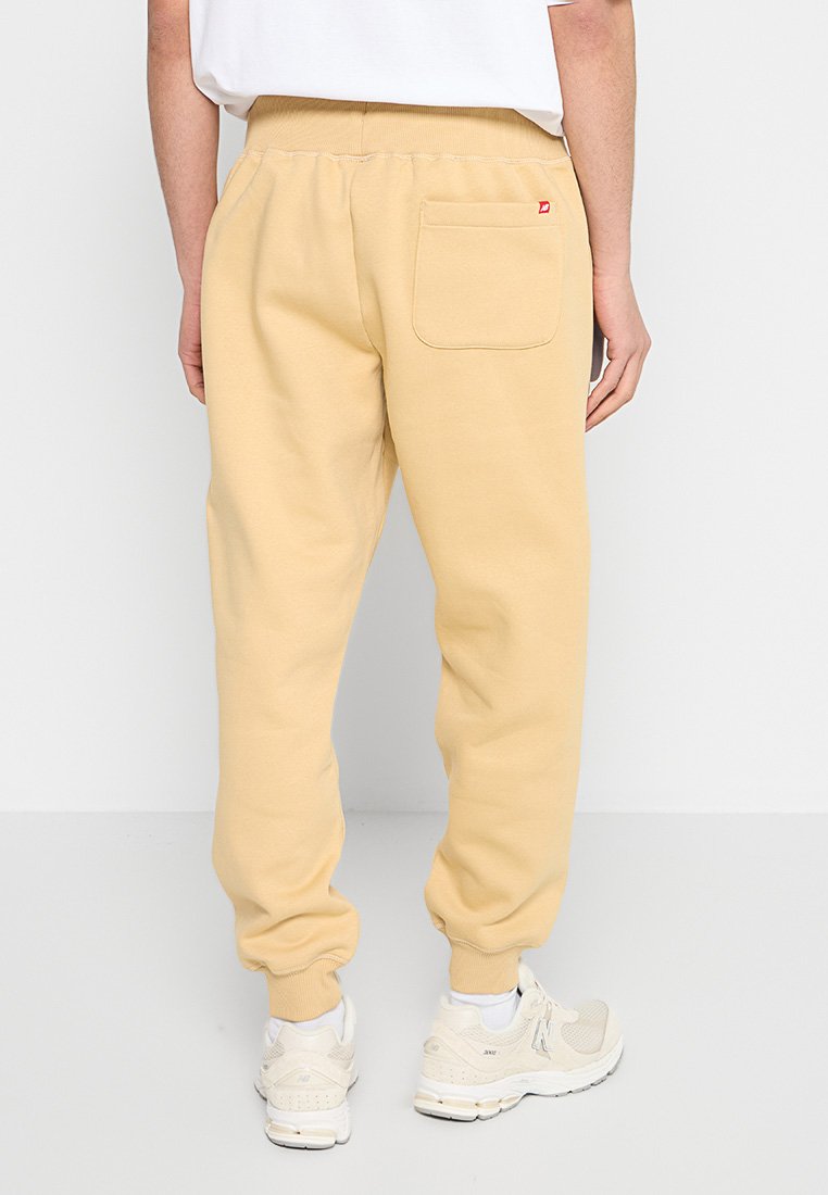 Pantalones de chándal beige hechos de tela suave, con una cinturilla elástica y un bolsillo trasero con parche. Combinados con zapatillas de color claro.