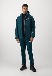 Veste imperméable turquoise avec une fermeture éclair à l'avant, accents noirs, poignets ajustables ; associée à un pantalon assorti et des chaussures noires.
