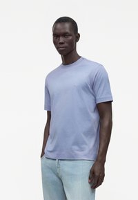 Hombre con camisa de manga corta azul claro y vaqueros de lavado claro, de pie con las manos en los bolsillos contra un fondo blanco.