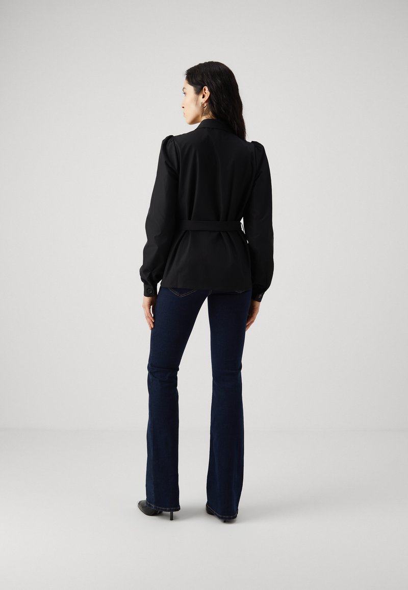 Schwarze Bluse mit langen Ärmeln und taillierter Passform, leicht gepufften Schultern und geknöpften Manschetten, kombiniert mit dunkelblauen Schlagjeans und schwarzen High Heels.