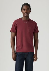 Bordeauxrote Baumwoll-T-Shirt mit Rundhalsausschnitt, kurzen Ärmeln und einer Brusttasche. Glatte Textur und minimalistisches Design, kombiniert mit dunklen Jeans.