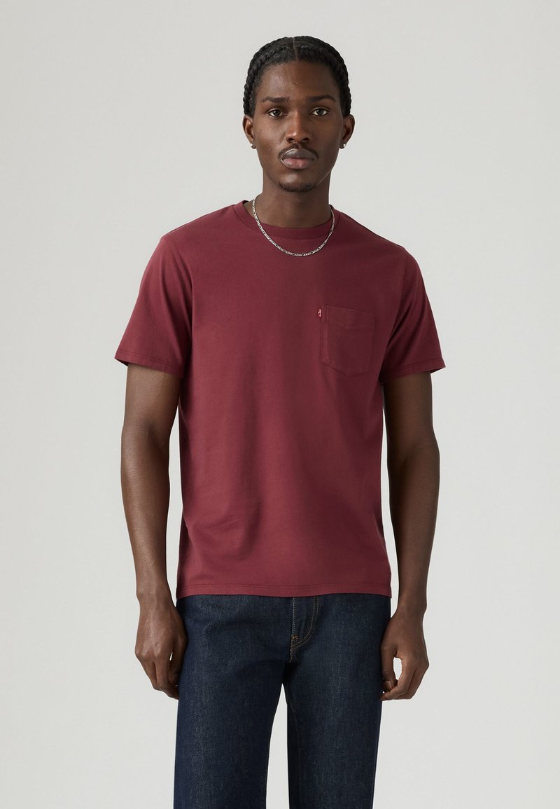 Bordeauxrote Baumwoll-T-Shirt mit Rundhalsausschnitt, kurzen Ärmeln und einer Brusttasche. Glatte Textur und minimalistisches Design, kombiniert mit dunklen Jeans.