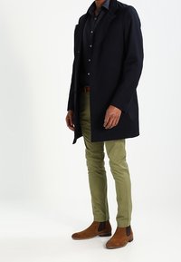 Black coat over a dark shirt, paired with olive-green pants and brown leather Chelsea boots. Smooth fabric and tailored fit.