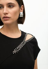 Black t-shirt with a rounded neckline, featuring a shoulder cutout and embellished with metallic rhinestones along the strap. Silver geometric earrings.