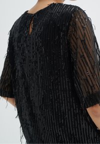 Abito nero realizzato in un tessuto testurizzato e ricoperto di paillettes. Maniche trasparenti e dettaglio a forma di occhio di bue sul retro, con frange che arricchiscono l'intero design.