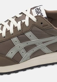 Sapatilha atlética castanha com detalhes em malha e couro, apresentando sobreposições verde-limão, atacadores brancos e uma sola de borracha texturizada.