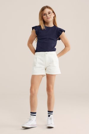 Jong meisje met lang haar, rond brilletje, marineblauw topje met ruches aan de mouwen, witte korte broek, gestreepte sokken en witte sneakers, staand met haar handen achter haar rug.