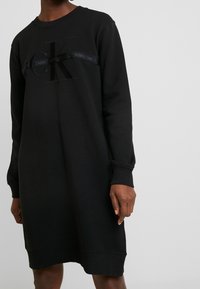 Musta college-mekko pitkillä hihoilla, jossa on pyöreä pääntie ja tekstuurilla varustettu logo rinnan kohdalla. Pehmeä kangas ja rento malli.