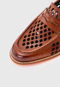 Καφέ δερμάτινο μοκασίνι με διάτρητες κυκλικές τρύπες και λεπτομερείς ραφές στο μπροστινό μέρος και το λουράκι, απομονωμένο σε φωτεινό φόντο.